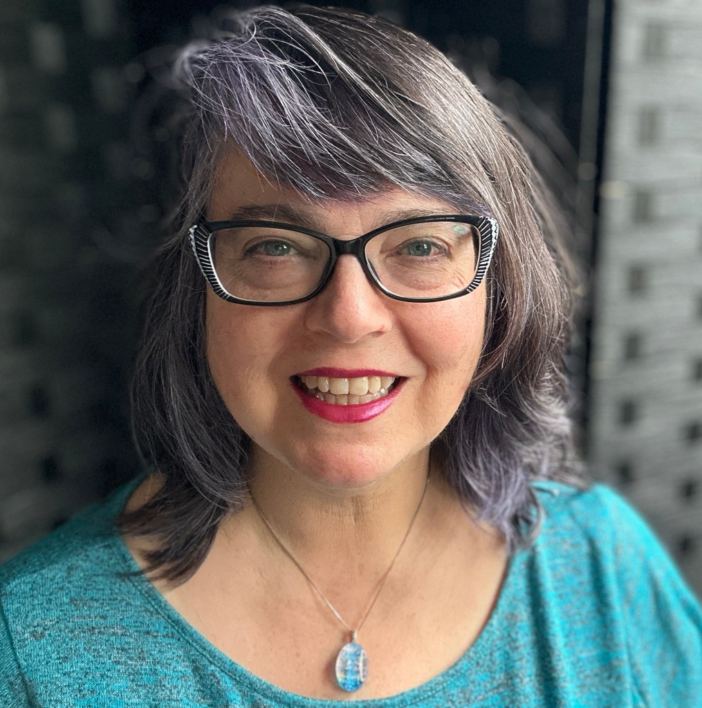 Izolda headshot. woman with dark gray background, turquoise blouse and necklace wearing glasses and cherry red lipstick